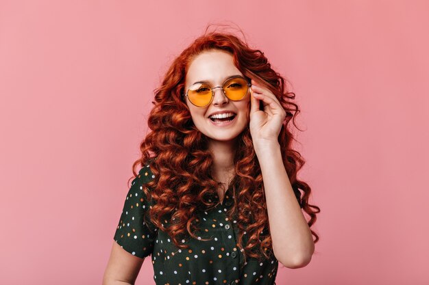 Beautiful ginger young woman posing in sunglasses with smile. Front view of laughing happy girl isolated on pink background.