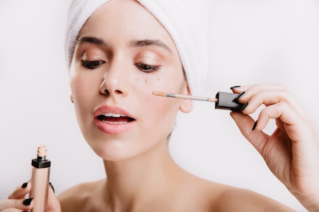 Beautiful girl after shower posing on white wall. Woman puts concealer to hide bruises under eyes.