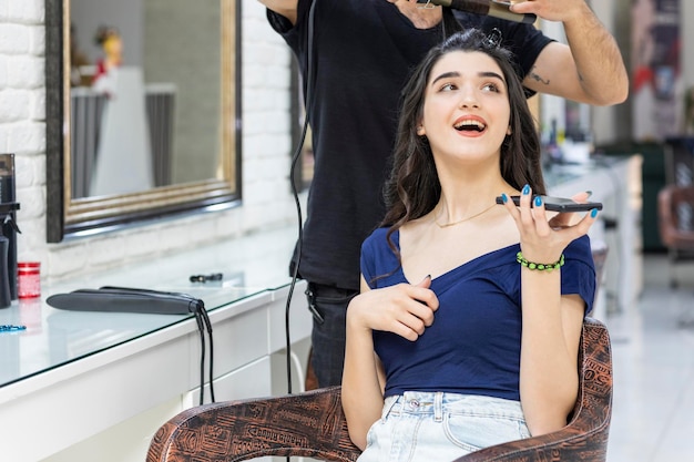 Beautiful girl at the barbershop holding phone and looking at side while Barber making her hair