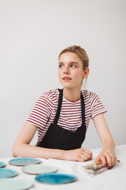 Beautiful girl in black apron and striped T-shirt sitting at the table with plates and pottery tools in hand thoughtfully looking aside at pottery studio