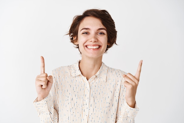 Beautiful girl in blouse pointing fingers up and smiling, standing against white wall in casual blouse