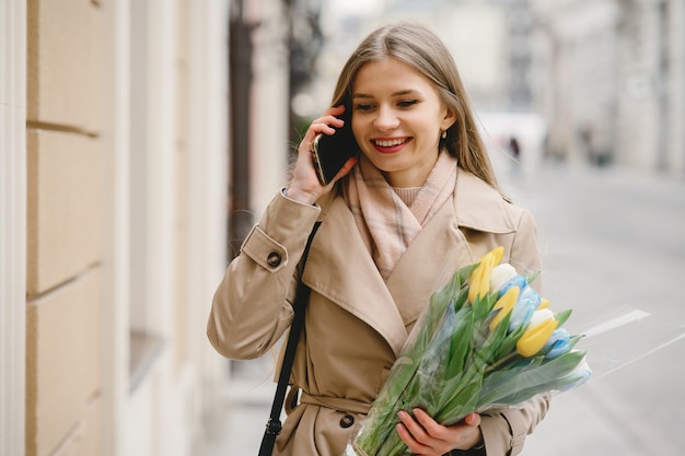 茶色のコートを着た美しい少女。春の街の女性。花の花束を持つ女性