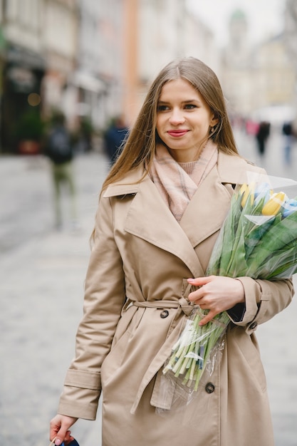 茶色のコートを着た美しい少女。春の街の女性。花の花束を持つ女性