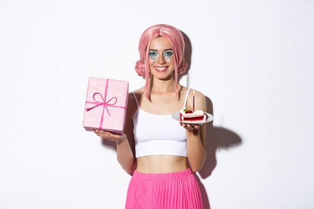 Beautiful girl celebrating birthday in pink wig, holding gift and b-day cake, standing.