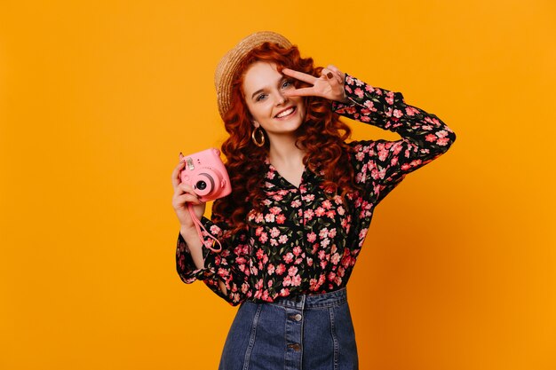 Beautiful girl in cheerful mood with snow-white smile shows sign of peace and makes photo on mini camera.