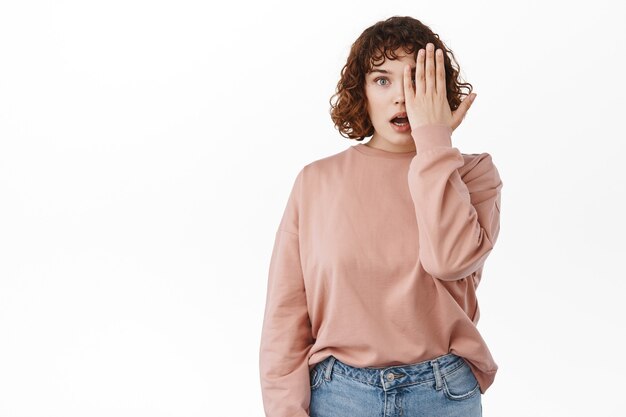 beautiful girl con half of face and look surprised, gasping and say wow amazed, staring at something awesome, standing on white.