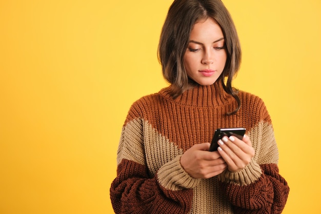 Beautiful girl in cozy sweater confidently using cellphone over yellow background