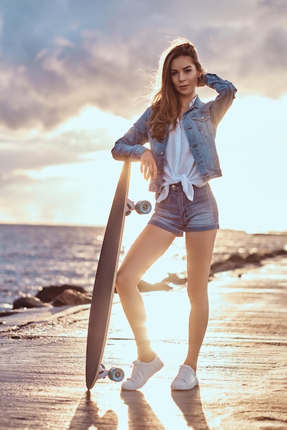 Beautiful girl dressed in denim shorts and jacket posing with a skateboard on the beach in cloudy weather during bright sunset.