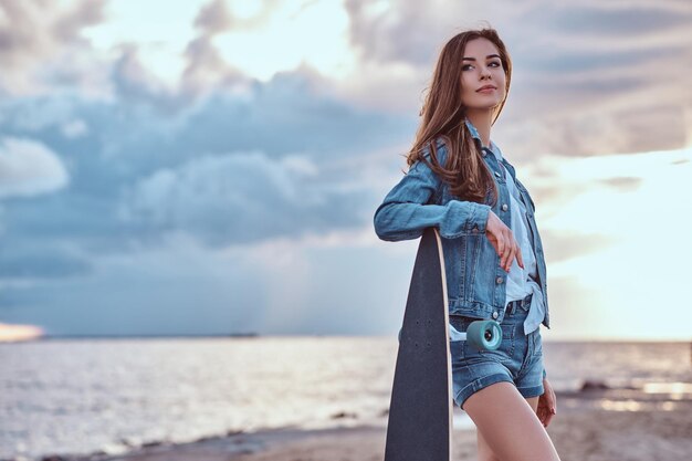 Beautiful girl dressed in denim shorts and jacket posing with a skateboard on the beach in cloudy weather during sunset.