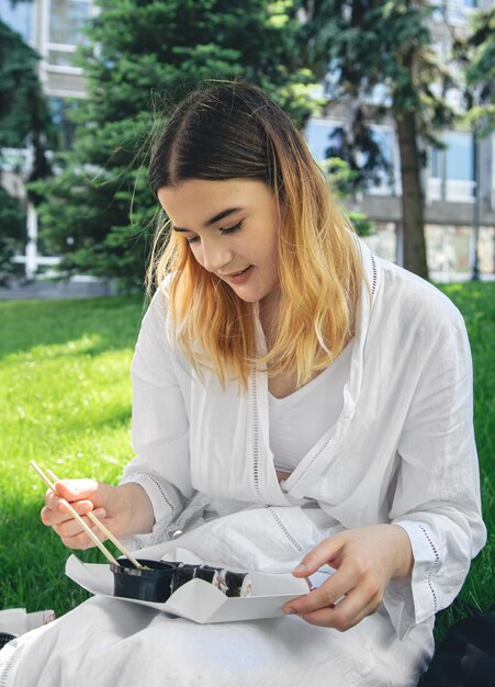 Красивая девушка ест суши, сидя на траве в парке