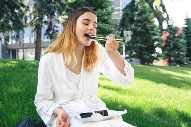 Beautiful girl eats sushi sitting on the grass in the park