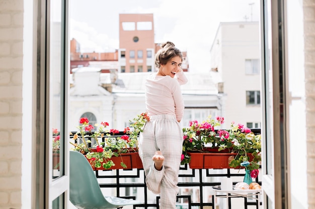 Beautiful girl from back in pajamas on balcony in sunny morning. She raises leg and looks excited to camera