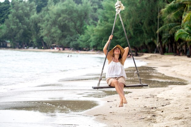 タイのビーチでぶら下がっているスイングの帽子で美しい少女