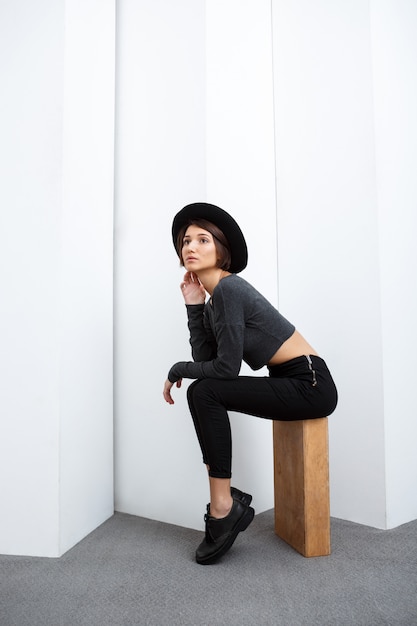 Beautiful girl in hat sitting on box over white wall