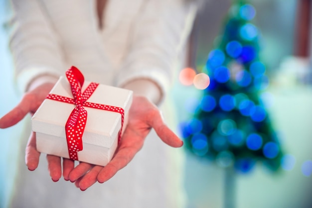 Beautiful girl holding a Christmas present in front of her. Happy woman in Santa hat standing near New Year tree and waiting for celebration.