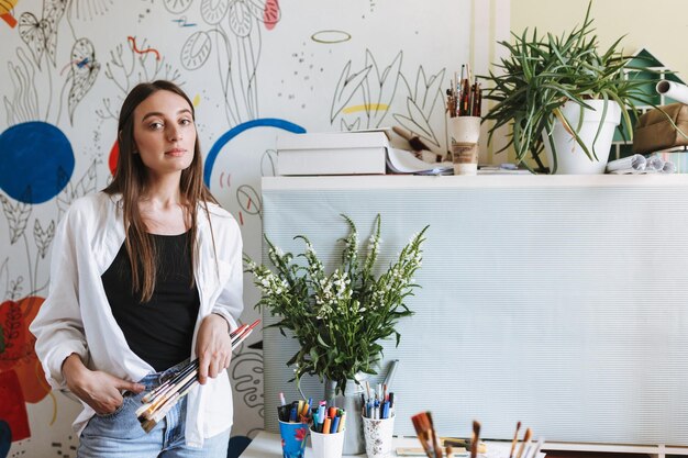 Beautiful girl holding paint brushes in hand while dreamily looking in camera with big patterns canvas on background at home