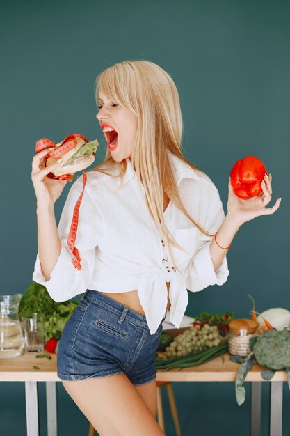 Beautiful girl make a salad. Sporty blonde in a kitchen. Woman chooses between burger and peper.