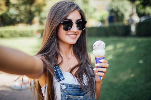 美しい少女は白い歯で自分撮り笑顔を作り、サングラスをかけてアイスクリームを保持します