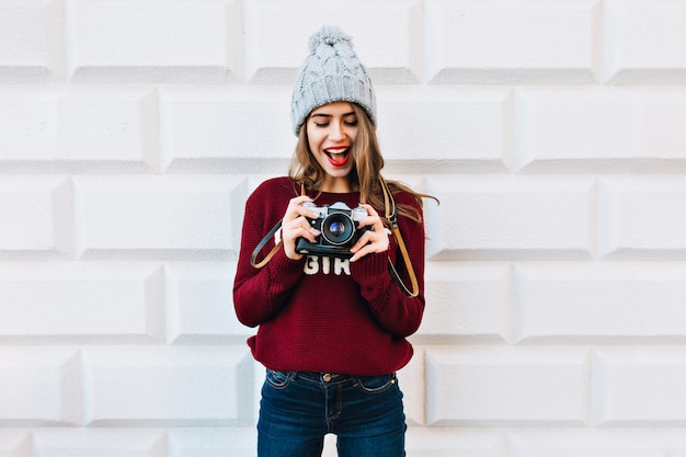 Beautiful girl in marsala sweater on grey wall . She wears knitted hat, is astonished looking at camera in hands.