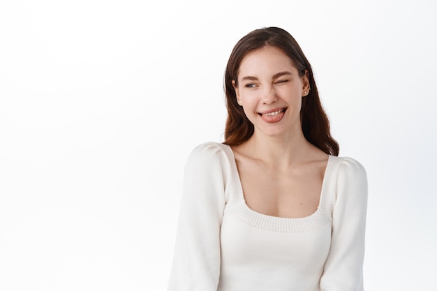 Beautiful girl model winking and showing tongue, making silly positive face, looking aside at company logo or promotional text, standing against white wall