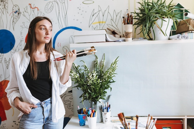 Beautiful girl near desk holding paint brushes in hand while thoughtfully looking aside with big patterns canvas on background at home