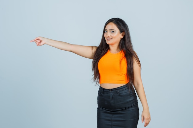 Free photo beautiful girl in orange top, skirt pointing to the side and looking cheerful , front view.