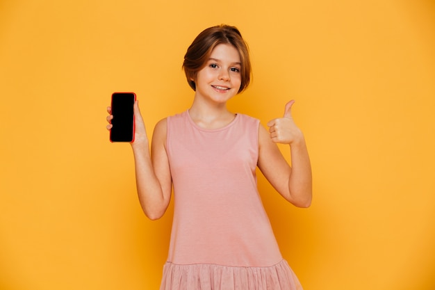 Beautiful girl in pink dress showing smartphone and thumb up isolated