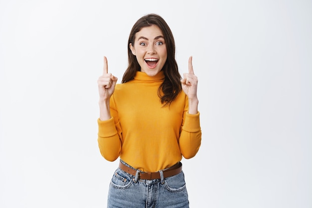 Beautiful girl pointing fingers up, advertising on white wall