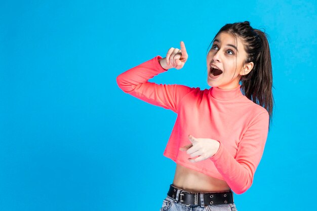 A beautiful girl pointing her finger in the air and standing on blue background High quality photo