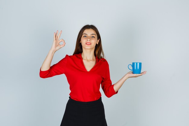 Beautiful girl in red blouse, black skirt holding cup, showing ok gesture and looking pleased , front view.