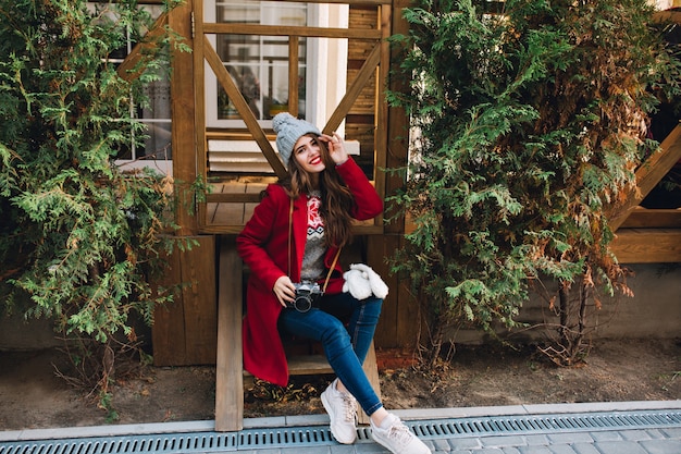 Foto gratuita bella ragazza in cappotto rosso e cappello lavorato a maglia che si siede sulle scale di legno. tiene la macchina fotografica sulle ginocchia e sorride.