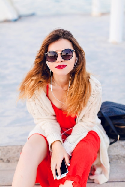 Free photo beautiful girl in red dress and white jacket sits on a pier, smiles, and listens to music on earphones on a smartphone.