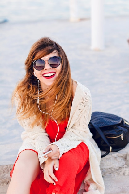 Beautiful girl in red dress and white jacket sits on a pier, smiles widely, and listens to music on earphones on a smartphone