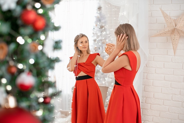 A beautiful girl in a red evening dress admires herself looking in the mirror. a young smiling woman prepares herself for the holiday. the blonde in the red dress.