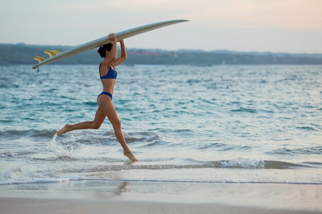 Красивая девушка бежит по пляжу с доской для серфинга.