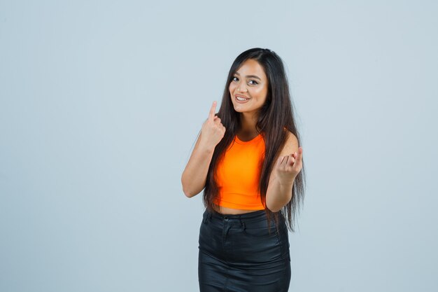 Beautiful girl showing number one with finger in orange top, skirt and looking pretty , front view.