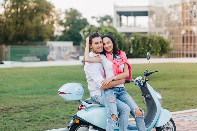 Beautiful girl in silver wristwatch relaxing with husband during warm spring day. Outdoor photo of laughing young man wears jeans posing on scooter, huggs with girlfriend.