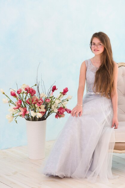 Красивая девушка сидит на кресле возле вазы с цветами, глядя на камеру