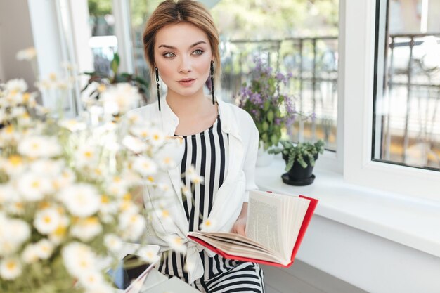 Beautiful girl sitting in coffee shop with book in hand and thoughtfully looking aside. Young lady with blond hair reading book in restaurant with bouquet of flowers on table