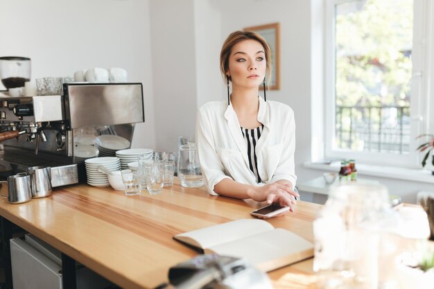 Beautiful girl sitting at the counter in coffee shop with mobile phone in hand and thoughtfully looking aside. Portrait of young pensive lady with blond hair leaning her elbow on counter in restaurant