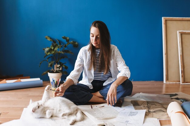 Beautiful girl sitting on floor with drawings dreamily playing with cute white cat while spending time at cozy home