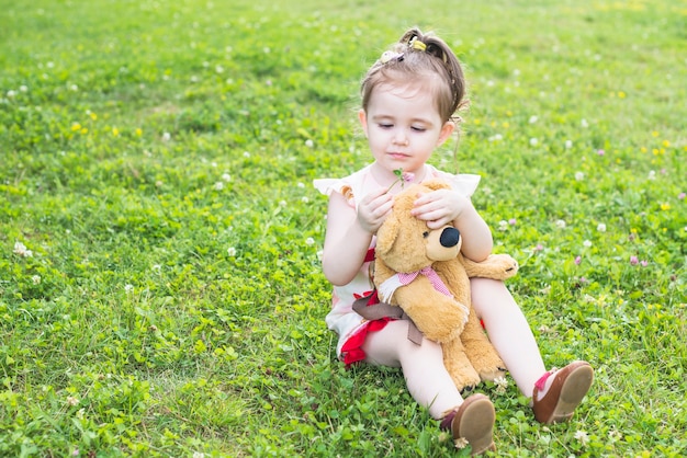 Beautiful girl sitting in the garden holding teddy bear looking at flower