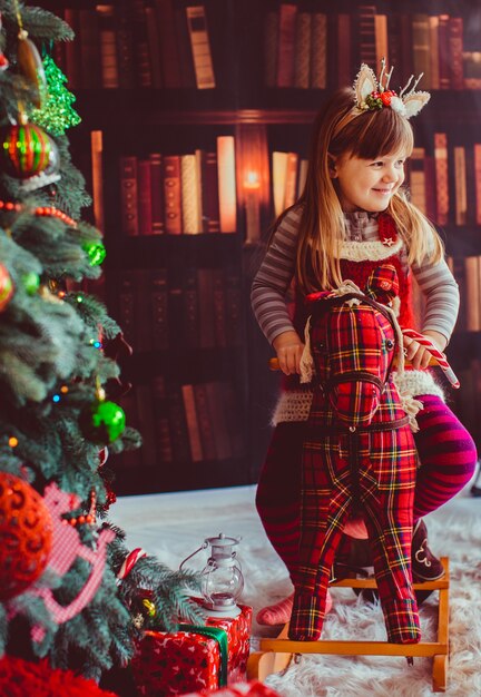 The beautiful girl sitting on the hobbyhorse near Christmas Tree