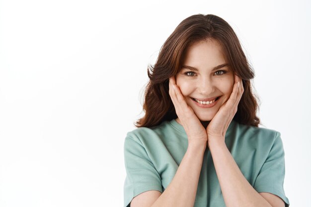 Beautiful girl smiling and blushing, touching her cheeks and looking flattered, receive compliments or praises, standing against white background