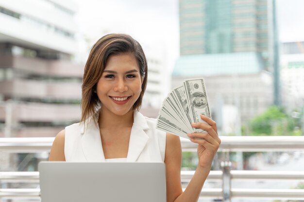 Beautiful girl smiling in business woman clothes using laptop computer and show money US dollar bills in hand