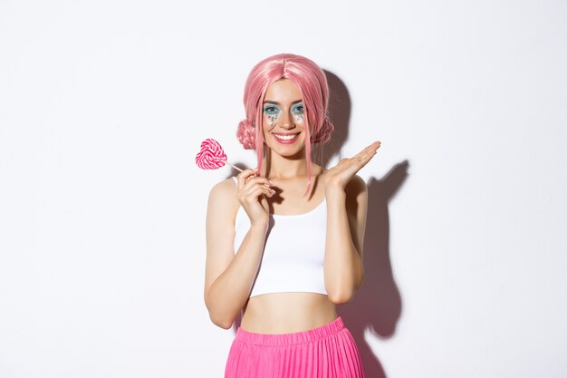 Beautiful girl smiling, wearing pink wig and fairy costume, holding heart-shaped candy, standing.