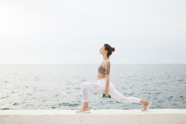 Beautiful girl in sporty top and white leggings doing exercises with dumbbells in hands while dreamily looking aside with sea on background