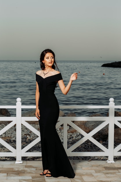 Foto gratuita bella ragazza che sta in vestito nero lungo dentro vicino alla spiaggia durante il giorno