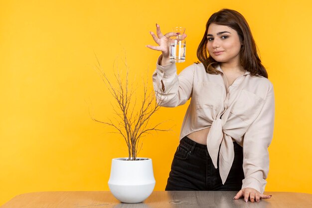 Beautiful girl standing on yellow background and holding glass of water High quality photo