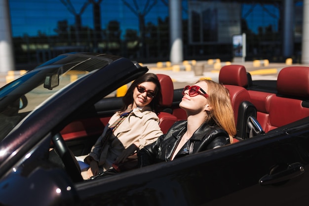 Bella ragazza in occhiali da sole che guida felicemente un'auto cabriolet con un amico sorridente vicino per le strade della città all'aperto